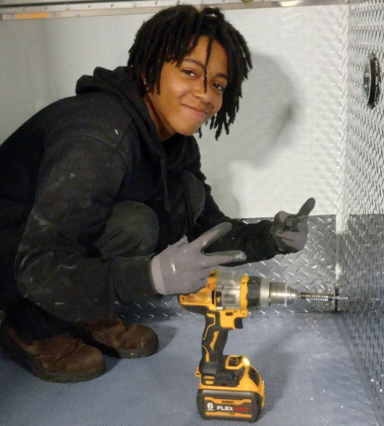 IVS Worker squatting in front of a metal sheet smiling and making peace signs towards the camera. A yellow drill sits infront of her.